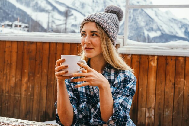 Beautiful sexy blond woman wearing knitting hat and holding hot drink mug outdoor on a wooden terrac