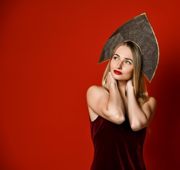 Beautiful sexy blonde Russian girl in traditional kokoshnik hat velvet festive dress on a red background