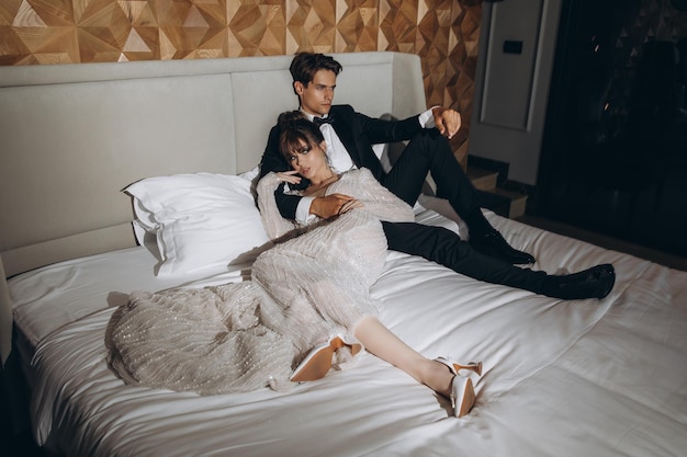 A beautiful and sexy bride in a wedding dress and a groom in a black suit in a luxury hotel on bed