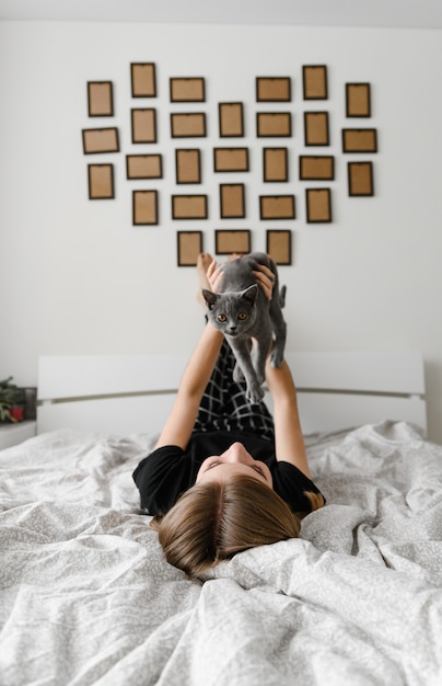 Красивая и сексуальная девушка лежит на кровати в самодельной пижаме, держа на руках счастливого шотландского серого котенка.