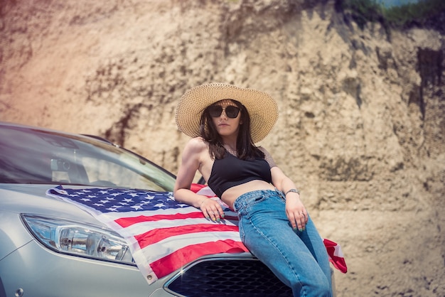 Bella ragazza sexy con bandiera americana e occhiali da sole vicino all'automobile nella cava di sabbia