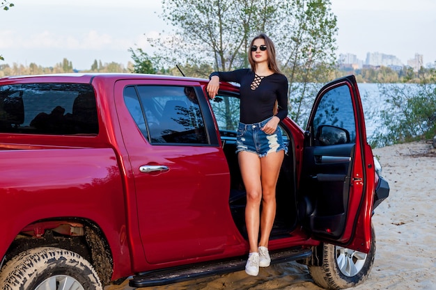 Beautiful sexy girl with long hair in short denim shorts and a black bodysuit posing near the open door of a red pickup truck. Girl posing near the car