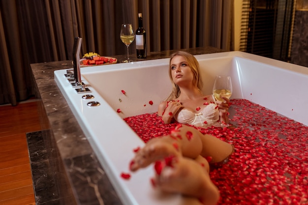 Beautiful sexy young blonde lying in bath tub full of water and many fresh red petals from rose flower drinking white wine