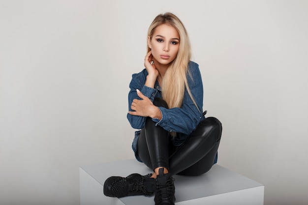 Beautiful sexy young girl with a hairstyle in fashion jeans clothes with black sneakers sits in studio