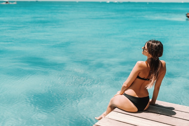 Beautiful shaped brunette in swimsuit an with sunglasses sitting on pier and enjoying sunny weather and ocean. Back turned.