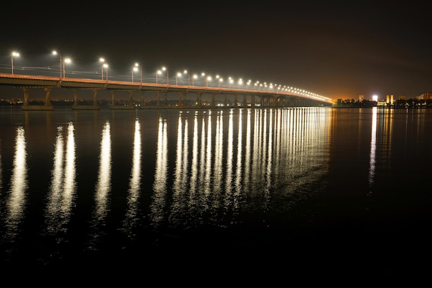 Beautiful shiny reflection of bright lanterns with cold light in the big Dnieper river under a long bridge passing over it