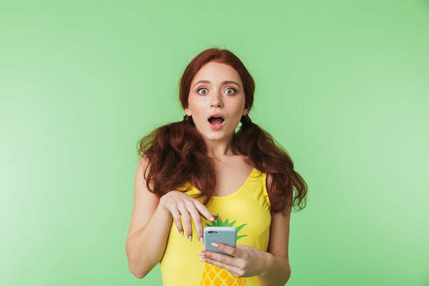 beautiful shocked emotional young redhead girl posing isolated over green wall background using mobile phone.