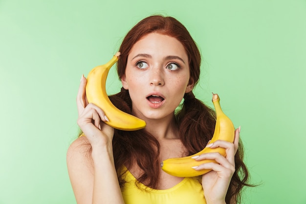 beautiful shocked excited young redhead girl posing isolated over green wall background with bananas fruits.