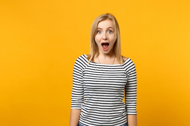 Beautiful shocked young blonde woman in striped clothes looking camera isolated on bright yellow orange wall background, studio portrait. People sincere emotions lifestyle concept. Mock up copy space.