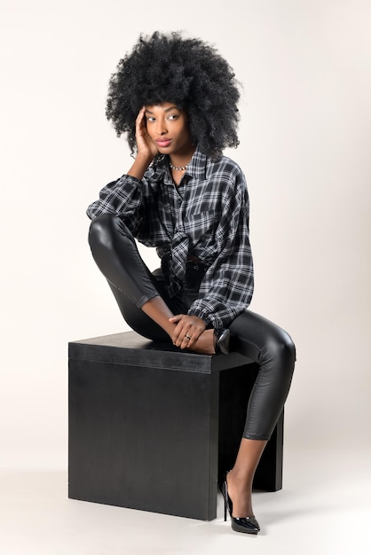 スタジオポートレートの黒い木製のスツールに物思いにふける気分であごに手で座っているスタイリッシュな革のズボンとハイヒールの美しい細い黒人女性