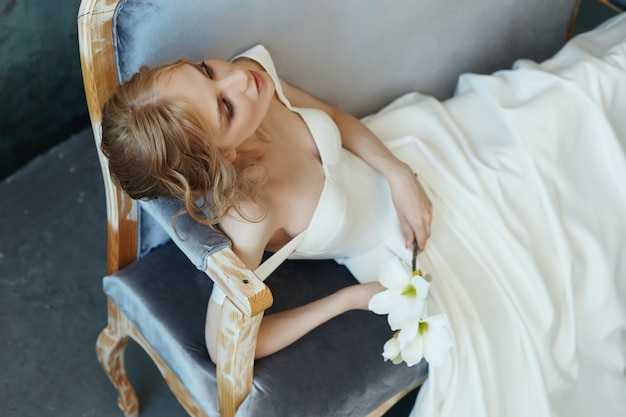 Beautiful slender blonde girl sitting on the sofa in a long white dress. Portrait of a woman with a flower in hand