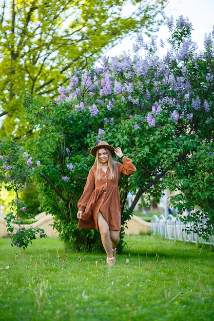Beautiful slender long-haired girl in a hat on a background of lilac flowers. She is of European appearance with a smile on her face. Young woman walks on a warm sunny day in a flowered park