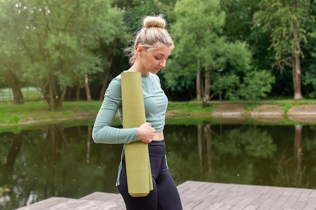 Beautiful slender woman holds green gym mat stand in the park in summer