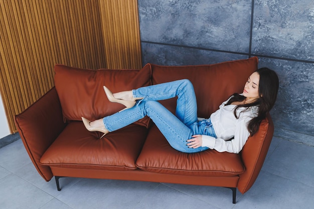 Beautiful slender woman legs in classic beige shoes with thin heels and jeans Stylish girl in heels sits on a brown leather sofa