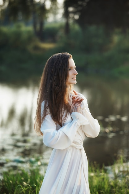 Bella donna snella in un lungo abito bianco cammina la mattina vicino al lago.