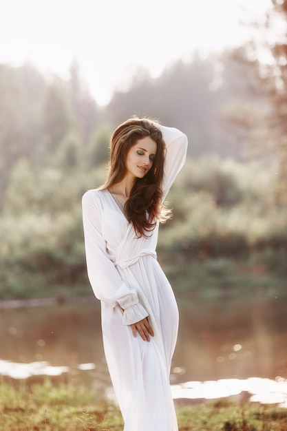 Photo beautiful slender woman in a long white dress walks in the morning near the lake