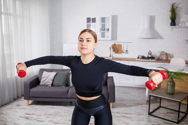 Beautiful slender young woman goes in for sports at home in a top and leggings. Fitness at home for a beautiful body. Exercises with dumbbells for the female body. Healthy lifestyle.