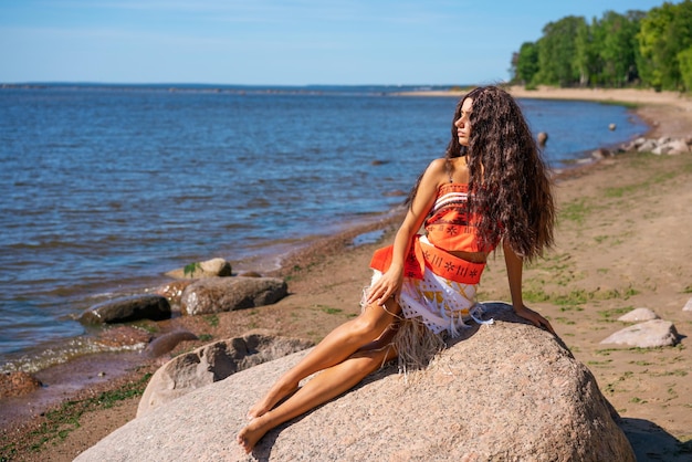 Beautiful slender young woman with wavy long hair sits on seashore on large