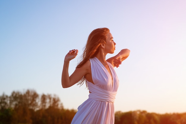 beautiful slender young womanwith long hair stands in a white dress in the wind near the sea at sunset