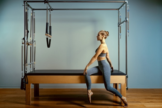 Beautiful slim fitness trainer on reformer gray background.