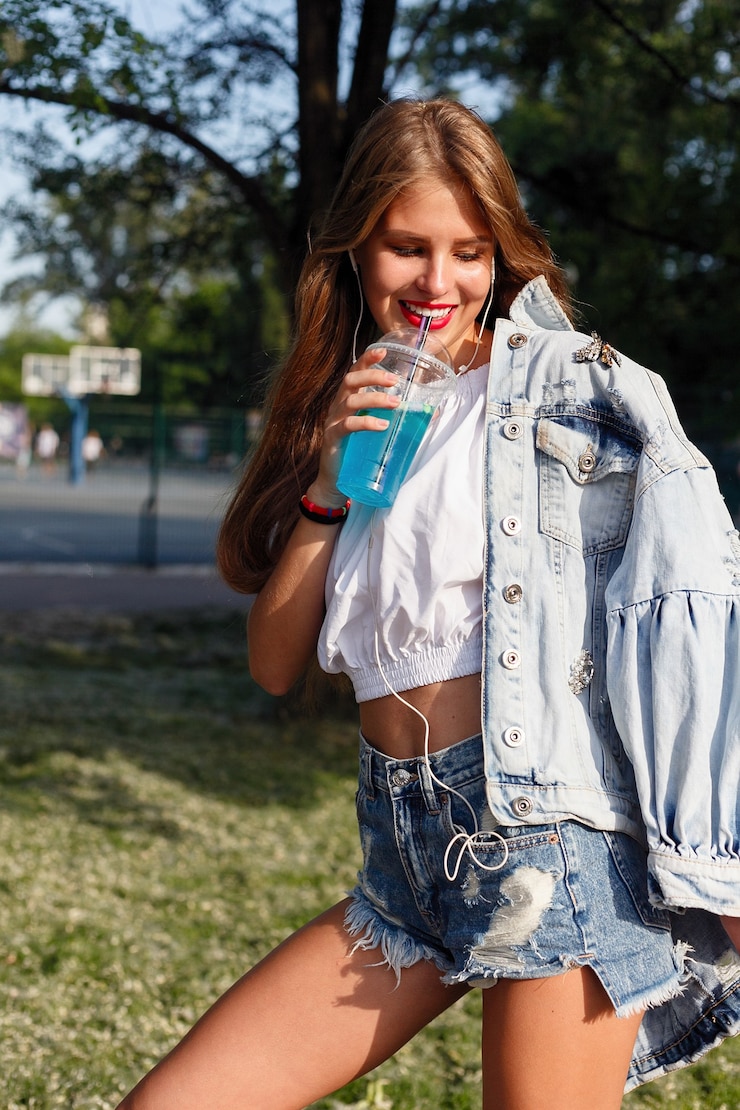 Photo beautiful slim girl drinking cocktail. adorable girl in a fashionable denim suit with long hairstyle and red lips drink refreshing lemonade lifestyle portrait of happy girl. she walks down the street