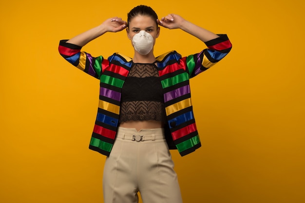 Beautiful slim girl fashion model posing in a protective respirator on a yellow space. LGBT community rainbow color jacket