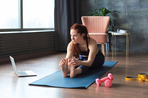 Beautiful slim sporty woman is stretching while sitting on the floor in the living room, watching in laptop where trainer showing exersices in zoom. Healthy lifestyle.