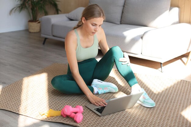 Beautiful slim sporty woman in sportswear is sitting on the floor with dumbbells and is using a laptop at home in the living room. Healthy lifestyle. Stay at home activities.