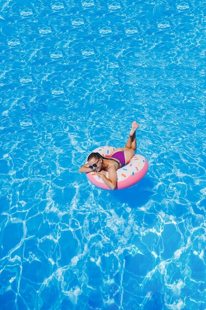 Beautiful slim young woman in a swimsuit enjoying the water park floating in an inflatable big ring on a sparkling blue pool smiling at the camera Summer vacation