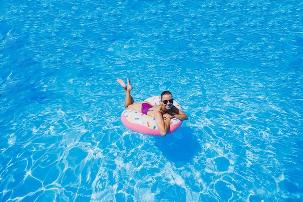 カメラに微笑んで輝く青いプールの膨脹可能な大きなリングに浮かぶウォーターパークを楽しんでいる水着の美しいスリムな若い女性夏休み