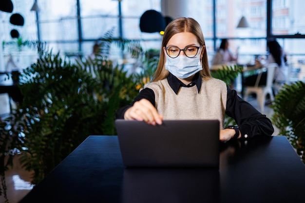 Photo a beautiful smart businesswoman in a large company holds an online meeting with business partners in a protective mask during the coronavirus, she sits at a table at a workplace with a laptop