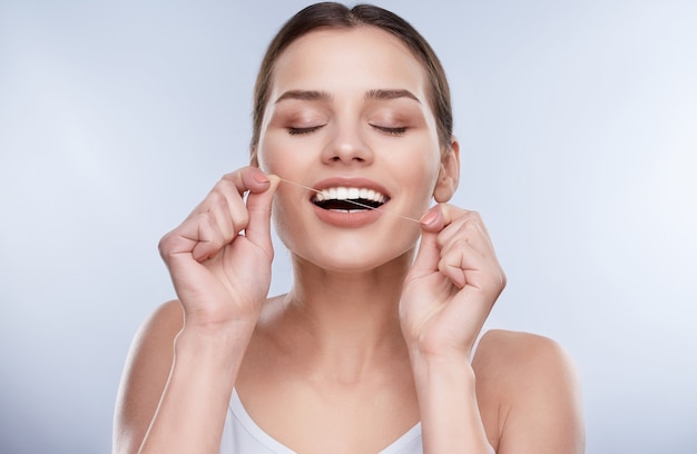 Beautiful smile, white strong teeth. Head and shoulders of woman with snow-white smile holding tooth thread, stomatological concept. Girl cleaning tooth with thread, closed eyes