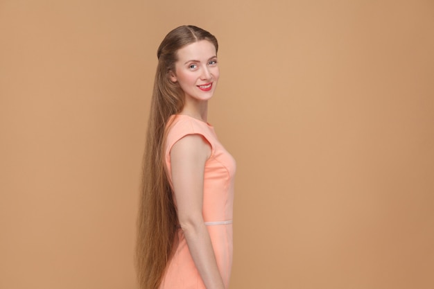 Beautiful smiley woman with long brown hair looking at camera. portrait of emotional cute pretty woman with makeup and long hair in pink dress. studio shot isolated on light brown or beige background