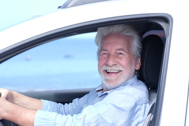 beautiful smiling adult whitehaired senior man sitting inside the car touching the steering wheel looking at camera elderly grandparent with white beard enjoying travel horizon over water