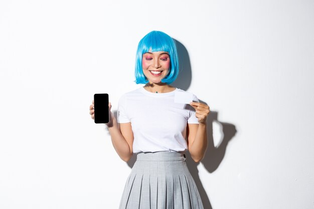 Beautiful smiling asian girl in blue wig looking pleased at credit card, showing smartphone screen, standing.