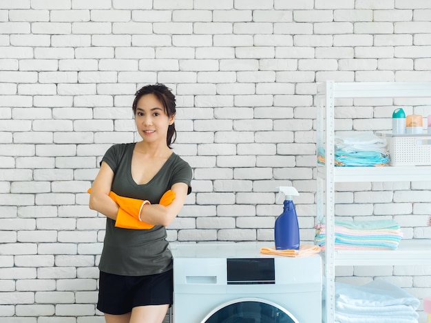 Красивая улыбающаяся азиатская женщина, счастливая домохозяйка в оранжевых защитных резиновых перчатках, стоя со сложенными руками рядом со стиральной машиной на белой кирпичной стене
