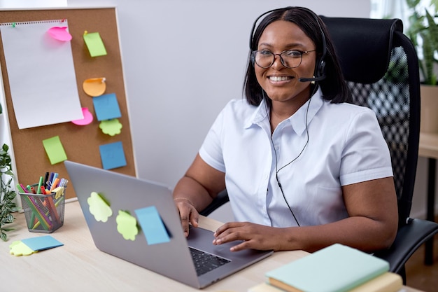 Beautiful smiling black female dispatcher with microphone working alone in office, using laptop. attractive young lady in formal wear enjoy being proficient. successful office worker. excited, happy