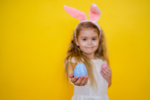 Beautiful smiling blonde girl with bunny ears holding out an easter egg in her hands selective focus on egg on a yellow background kid celebrate easter High quality photo