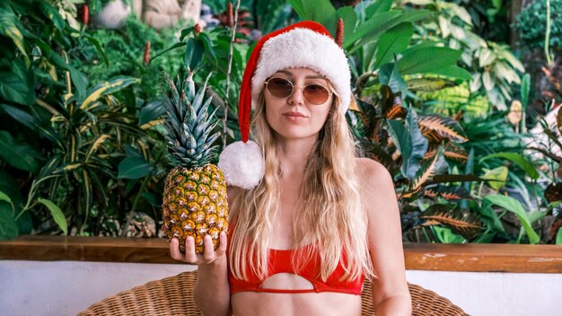 Foto bella bionda sorridente in cappello di babbo natale che esamina la macchina fotografica che tiene ananas indossando di nuovo gli occhiali da sole