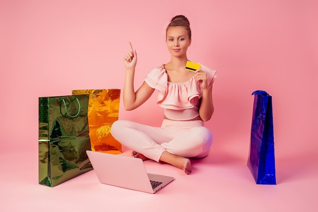 Beautiful smiling blonde woman shopaholic bag and credit card in her hand sitting on floor with laptop pink background in studio . concept of seasonal black friday sale and online shopping