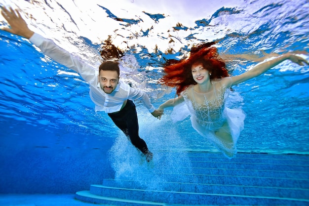 Beautiful smiling bride and groom in wedding dresses swim underwater in the pool on a sunny day