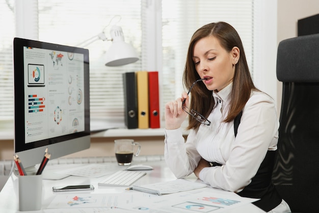 Beautiful smiling brown-hair business woman in suit and glasses sitting at the desk with cup of coffee, working at computer with documents in light office, looking paper