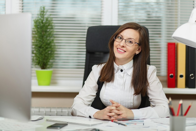 The beautiful smiling brown-hair business woman in suit and glasses sitting at the desk with tablet, working at computer with modern monitor with documents in light office, looking at the camera