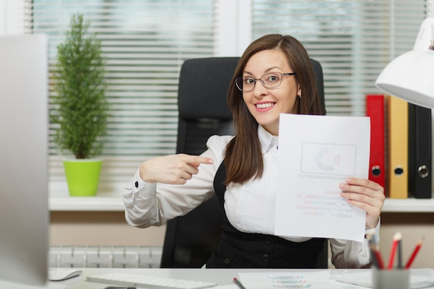 Beautiful smiling brown-hair business woman in suit and glasses sitting at the desk, working at computer with documents in light office, showing index finger on blank sheet. With place for text.