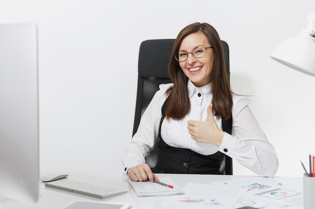 책상에 앉아 안경을 쓰고 웃고 있는 아름다운 갈색 머리 비즈니스 여성, 밝은 사무실에 있는 문서가 있는 현대적인 모니터로 컴퓨터 작업, 엄지손가락을 보여주는