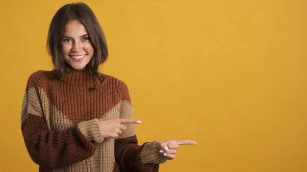Красивая улыбающаяся брюнетка в уютном вязаном свитере счастливо показывает указательные пальцы в сторону на красочном фоне