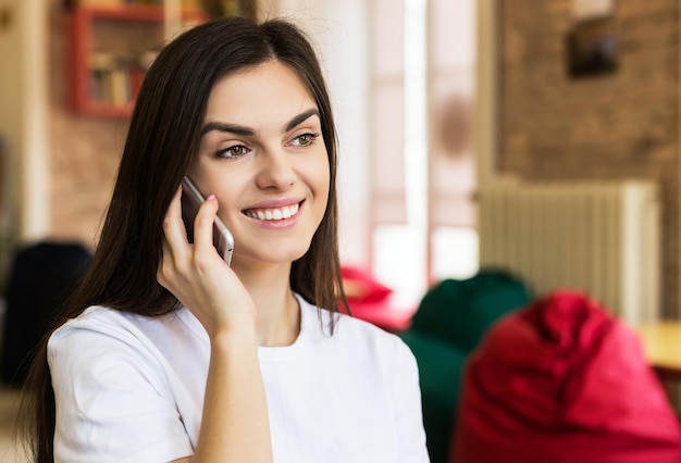 美しい笑顔のブルネットの女の子は、スタイリッシュでモダンなホームルームで話す喜びのためにスマートフォンを使用しています