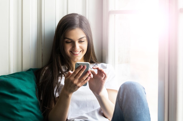 Beautiful smiling brunette girl wears white tshirt using smartphone sitting on the windwsill