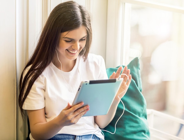 Beautiful smiling brunette girl wears white tshirt using tablet computer for videochat sitting on the windwsill