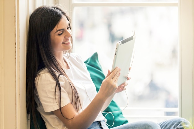 美しい笑顔のブルネットの少女は、風車に座っているビデオチャットのためにタブレットコンピューターを使用して白いTシャツを着ています